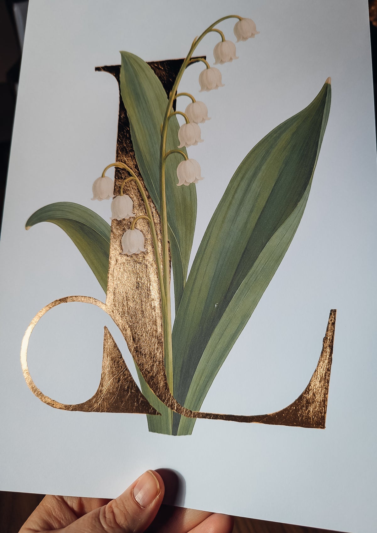 A hand holds a white art print featuring a large decorative letter L decorated with lily of the valley flowers and green leaves, with metallic gold foil accents.