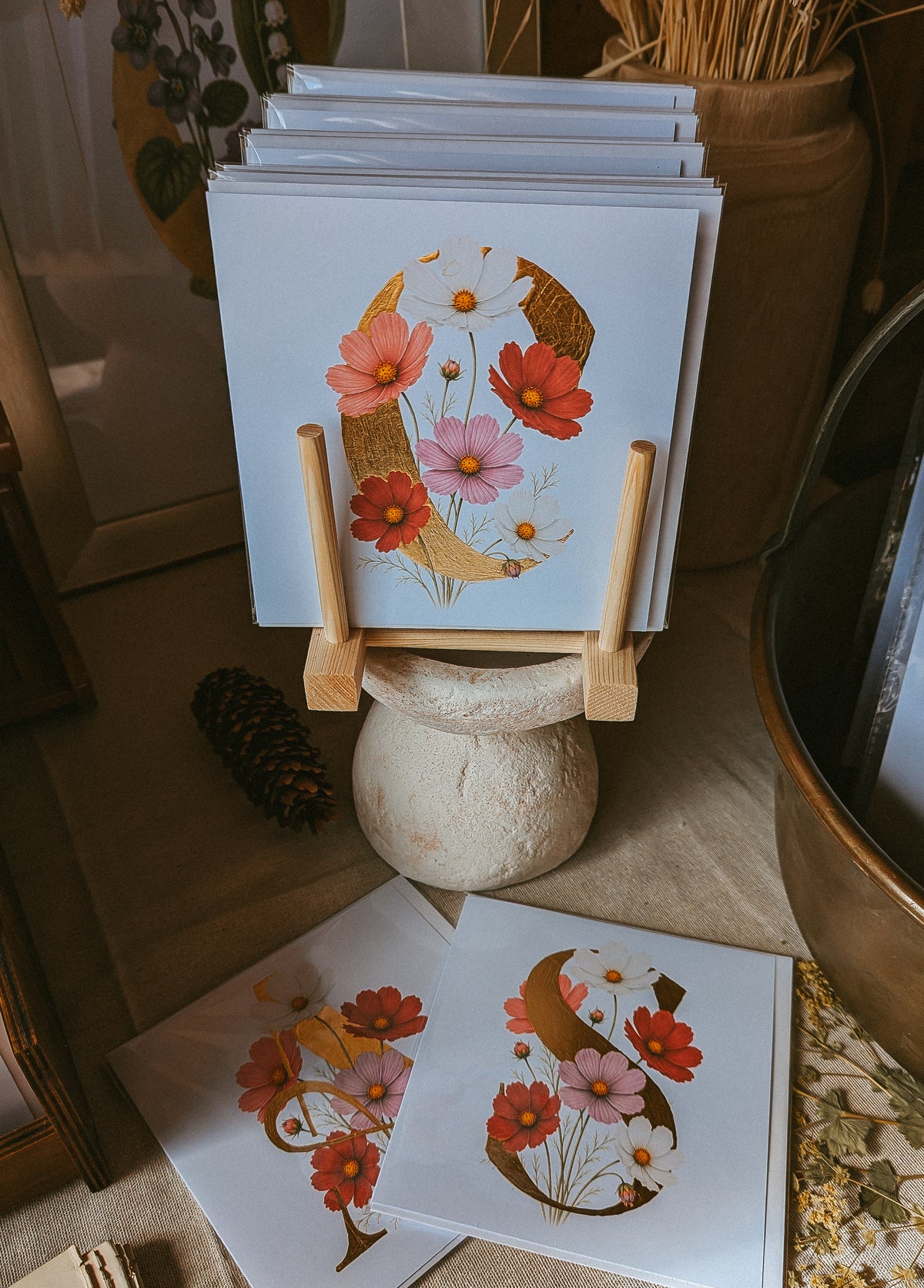 A stack of art cards displayed on a small wooden easel stand, with two prints laid out in front on a tabletop. The prints feature a stylised letter 'C' decorated with red, pink, and white Cosmos flowers and gold leaf-like accents, set in a rustic, warm-toned room.