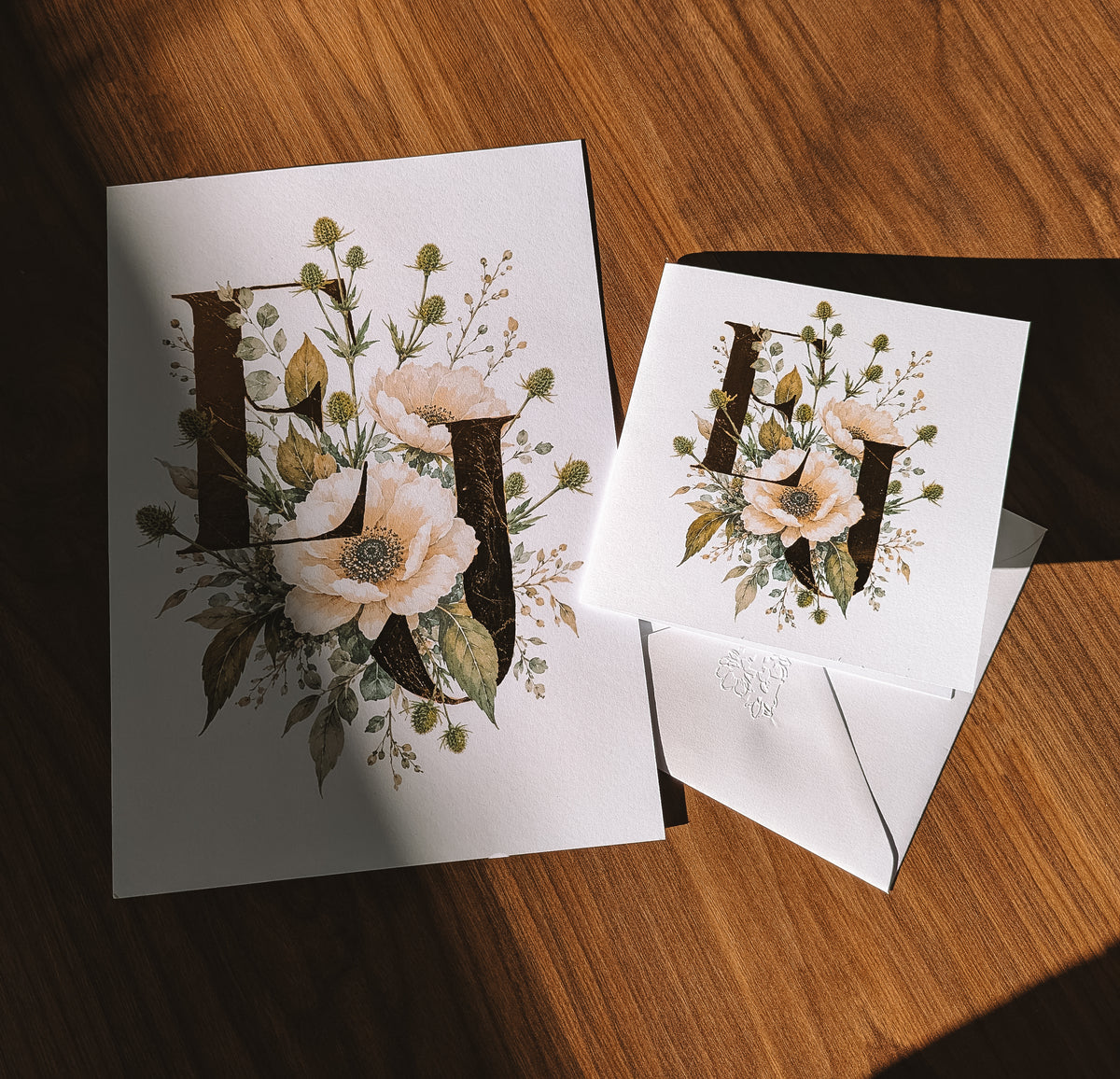 A styled flat-lay of a white stationery card featuring a large decorative monogram letters E & J surrounded by delicate floral illustrations, shown alongside a matching white envelope on a warm wooden surface.