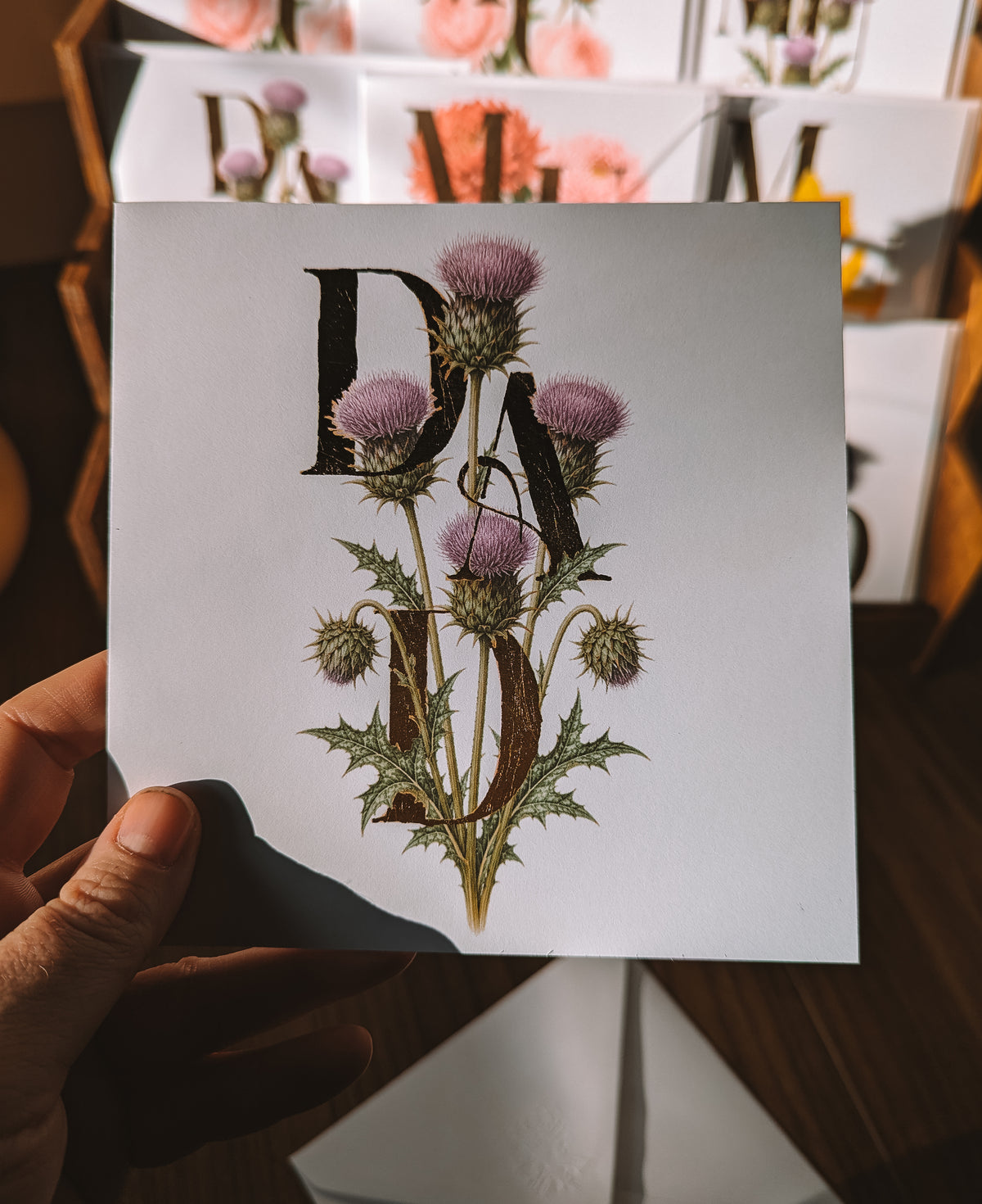 A hand holds a square white card featuring a vintage-style botanical illustration of purple thistle flowers and green leaves, overlaid with large gold foil serif letters spelling 'DAD'. The background shows a blurred wooden shelf with similar floral prints.