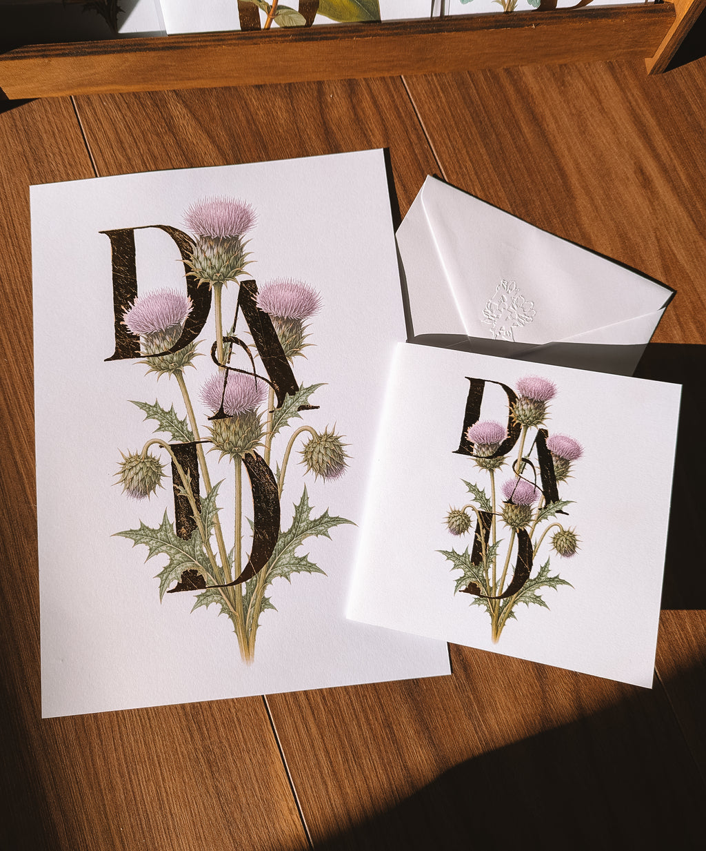 White greeting card and print on a wooden table, featuring a botanical design with purple thistle flowers and large decorative gold foil letters spelling 'DAD'.