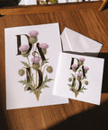 White greeting card and print on a wooden table, featuring a botanical design with purple thistle flowers and large decorative gold foil letters spelling 'DAD'.