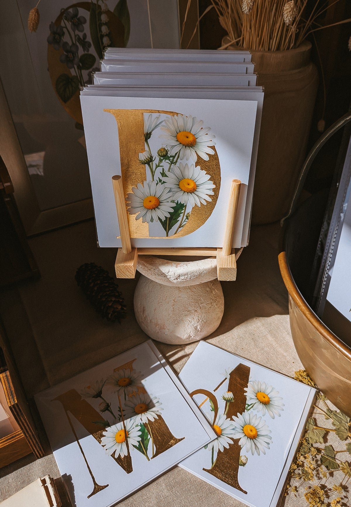 A stack of greeting cards displayed on a small wooden stand in a styled interior setting. The front card features a large gold letter 'D' with white daisies that have yellow centers.