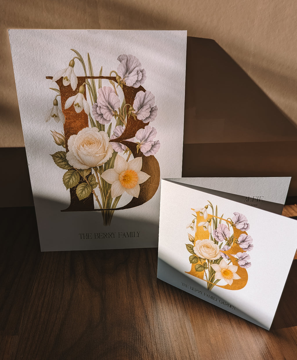 Botanical Print and card displayed on a wooden surface against a tan backdrop. The print shows a decorative floral illustration integrated into the letter B, with the text 'THE BERRY FAMILY' below.