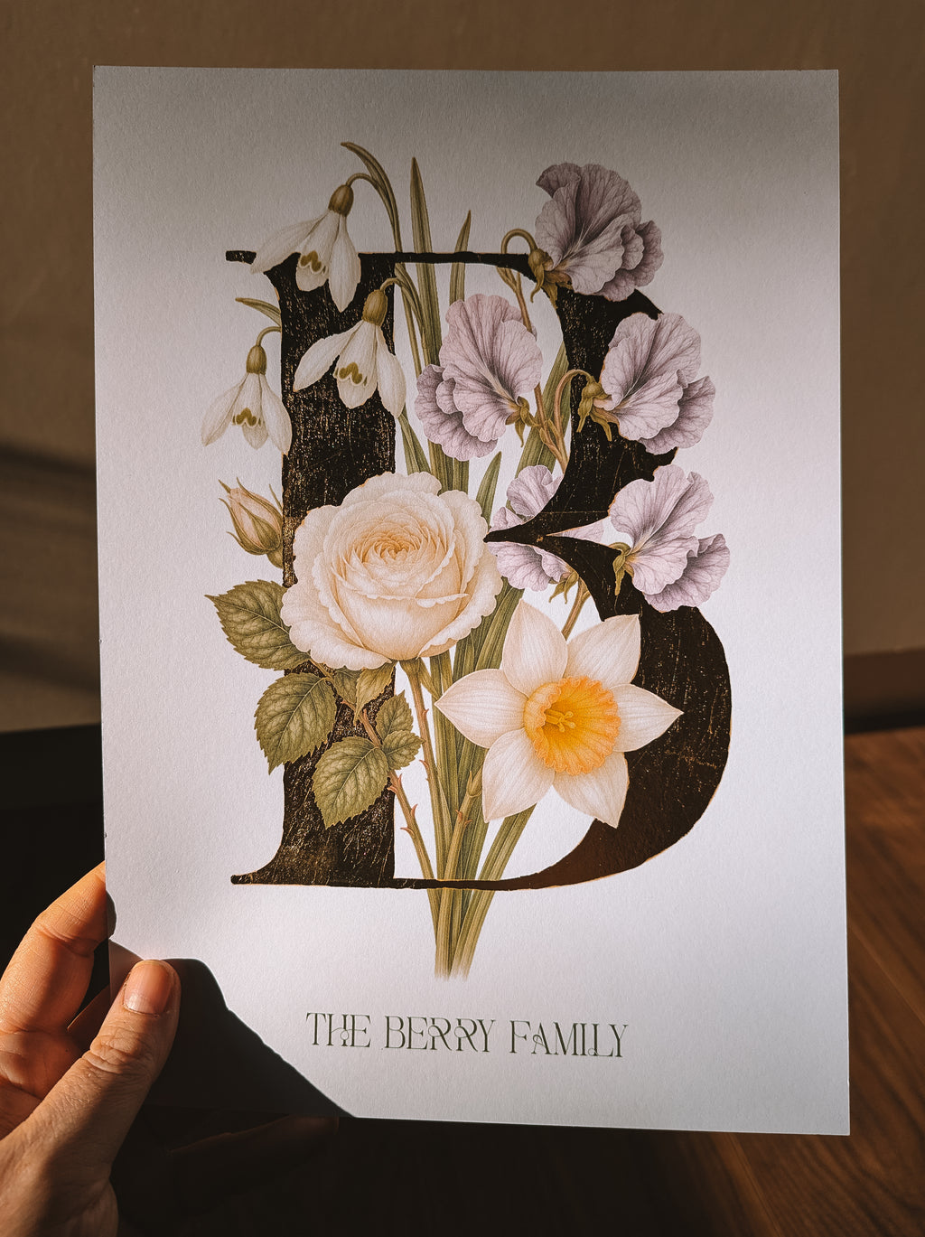 A hand holds a white art print featuring a large decorative letter B filled with illustrated flowers, including a pale pink sweet pea, white snowdrops, rose, and a daffodil, with the text 'THE BERRY FAMILY' below. The background shows a warm brown wooden surface and wall.