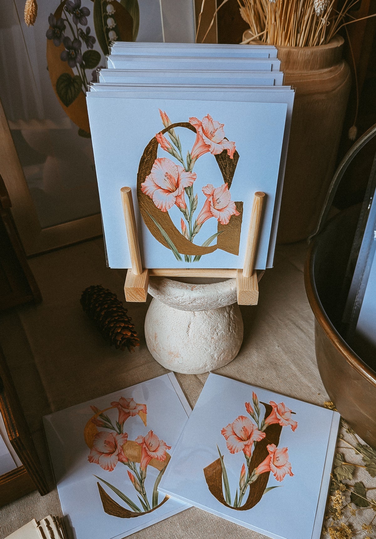 A stack of square art cards displayed on small wooden stands, with two prints laid flat in front. The prints feature a large decorative letter 'G' filled with pink Gladiolus flowers and green leaves on a pale blue background.