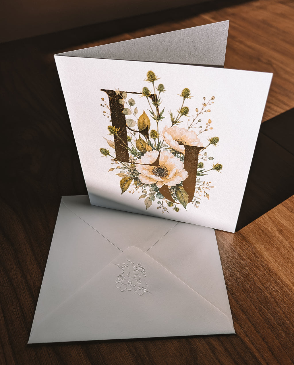A white greeting card with large decorative floral letters 'E&J' and an embossed floral envelope, displayed on a dark wood surface.