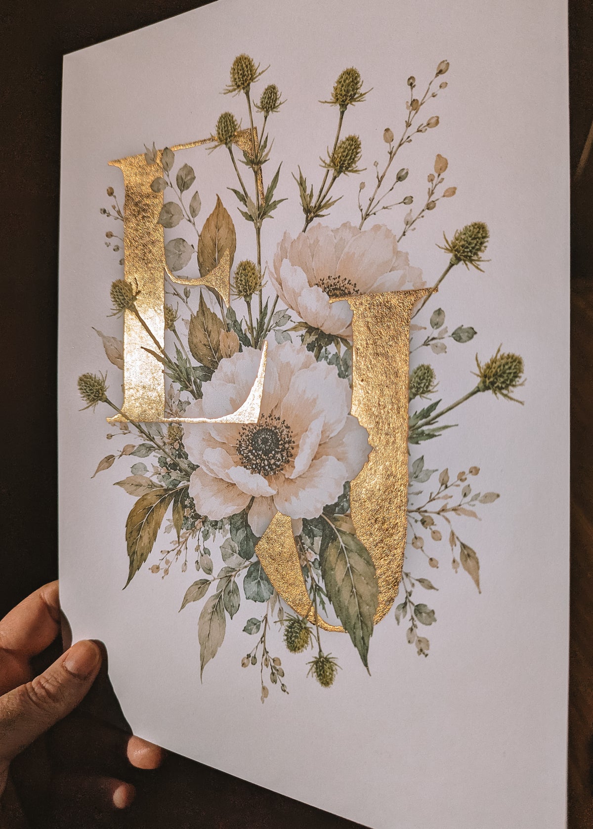 A hand holds a white art print featuring large decorative letters 'E&J' filled with white flowers, green leaves, and gold foil accents, set against a dark background.