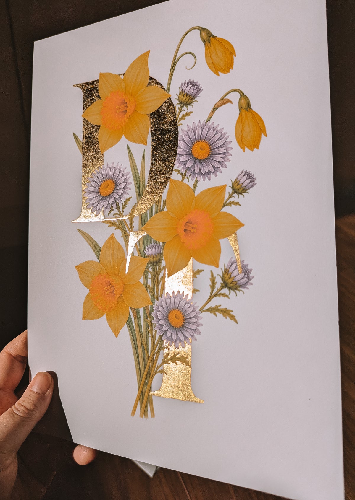 A hand holds a white art print featuring a large gold letters 'D&T' decorated with yellow daffodils and aster flowers, set against a dark wooden background.