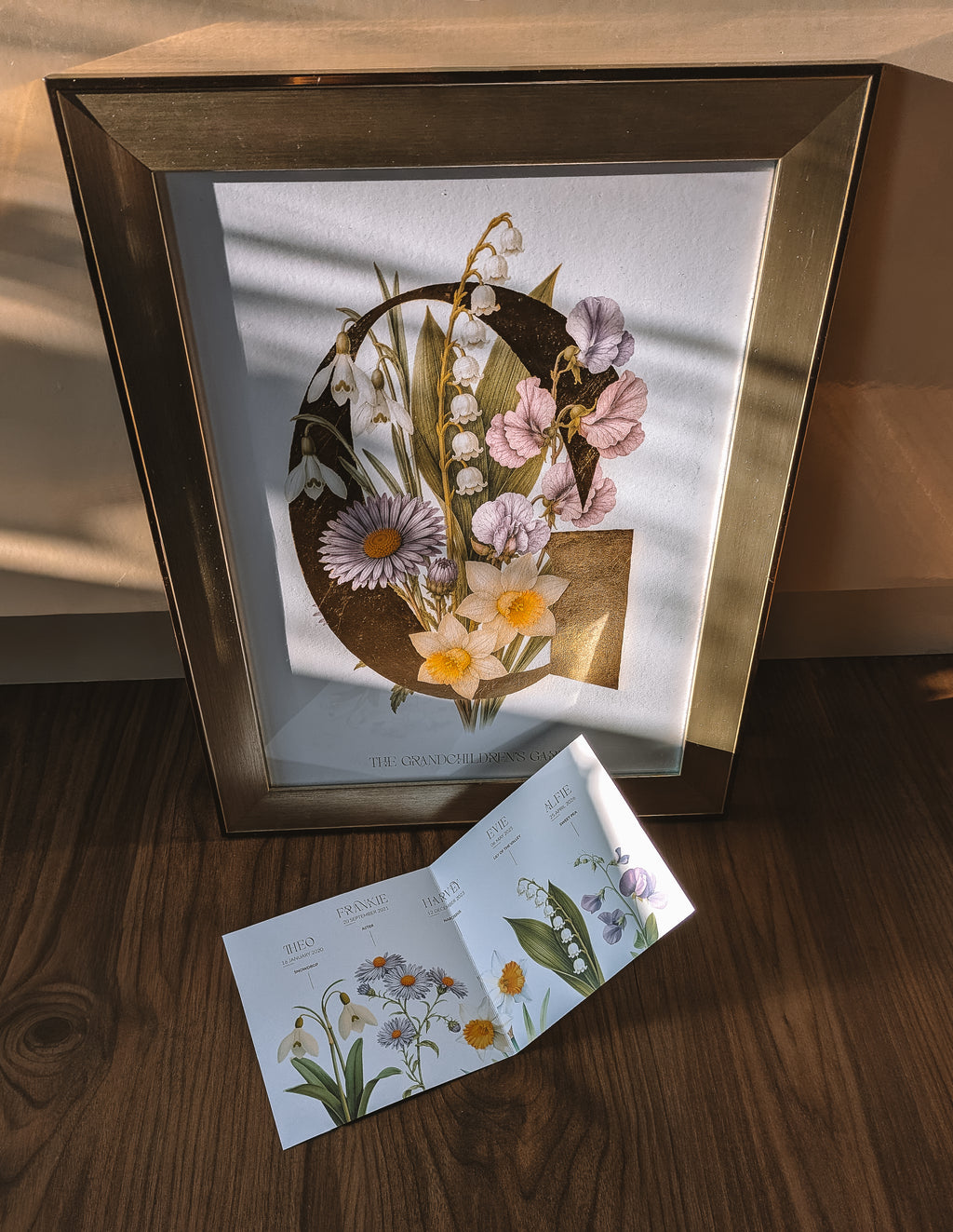 A framed botanical art print featuring a large gold letter 'G' adorned with flowers, including lily of the valley, asters, snowdrops, narcissus and sweet peas, set against a white background. The frame is gold, and the print is styled on a wooden surface with warm sunlight casting shadows.
