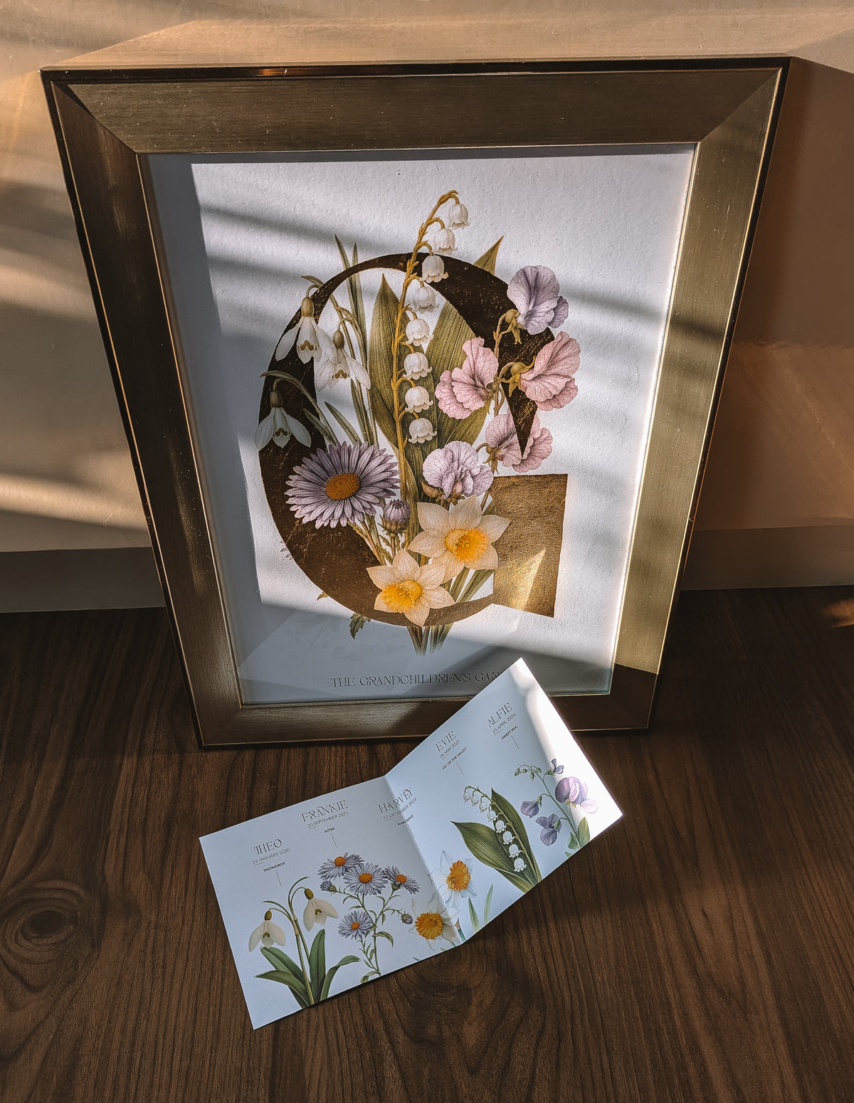 A framed botanical art print featuring a large gold letter 'G' adorned with flowers, including lily of the valley, asters, snowdrops, narcissus and sweet peas, set against a white background. The frame is gold, and the print is styled on a wooden surface with warm sunlight casting shadows.