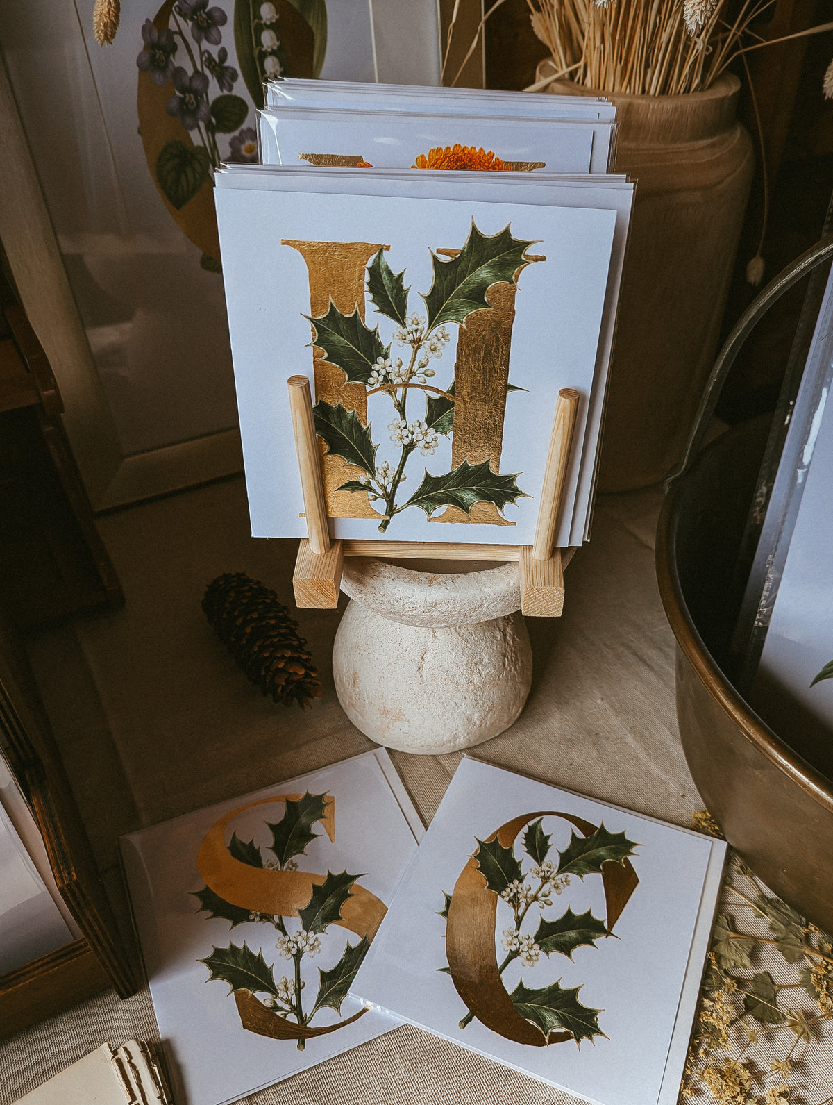 A stack of white greeting cards displayed on a small wooden stand, featuring large gold foil letters with green holly and small white berries. Two cards are placed in front on a beige fabric surface, with dried botanicals and rustic decor in the background.