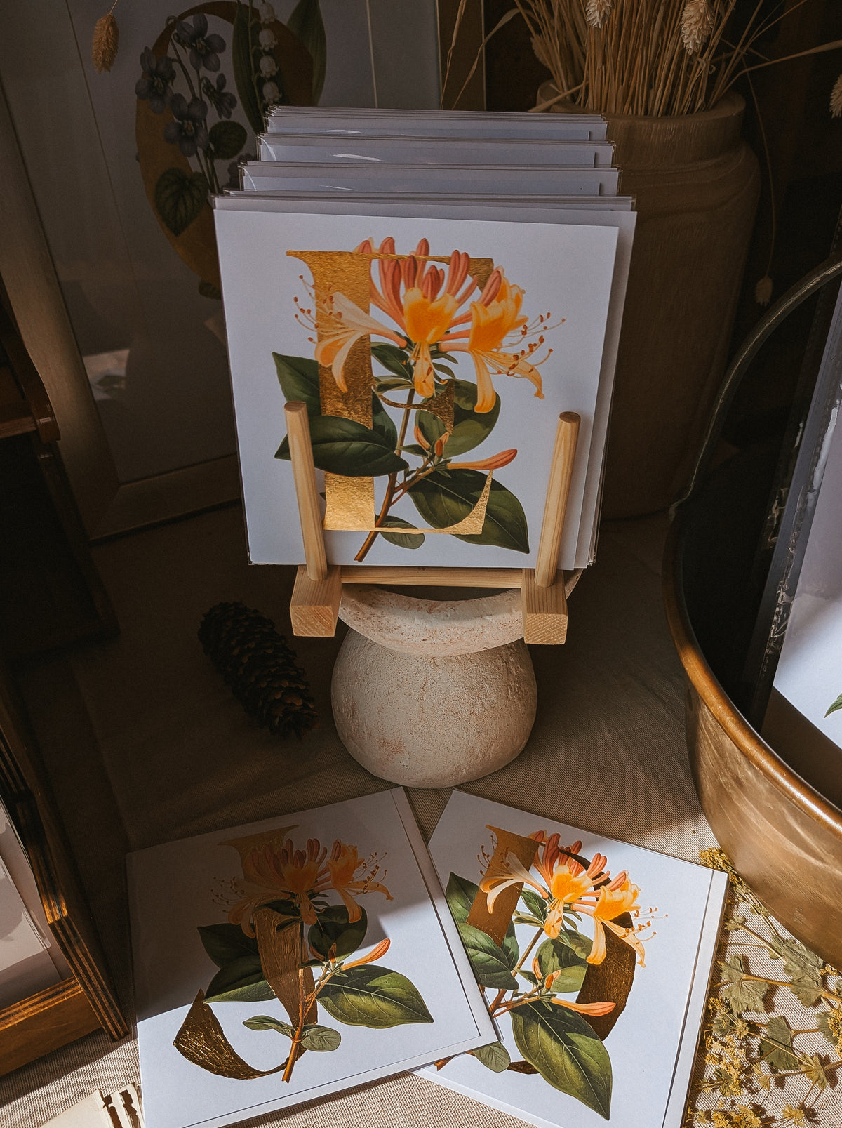 A stack of botanical art prints featuring Honeysuckle and green leaves, displayed on a small wooden easel stand with two prints laid out in front on a tabletop.