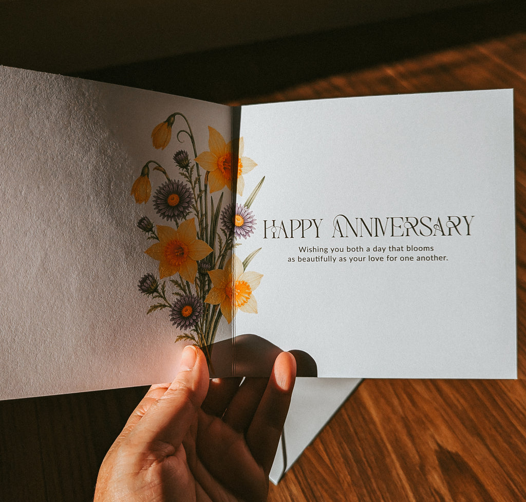 An open greeting card held in a hand, showing a floral illustration on the left page and the words “HAPPY ANNIVERSARY” with a short message on the right page, set against a warm wooden tabletop background.