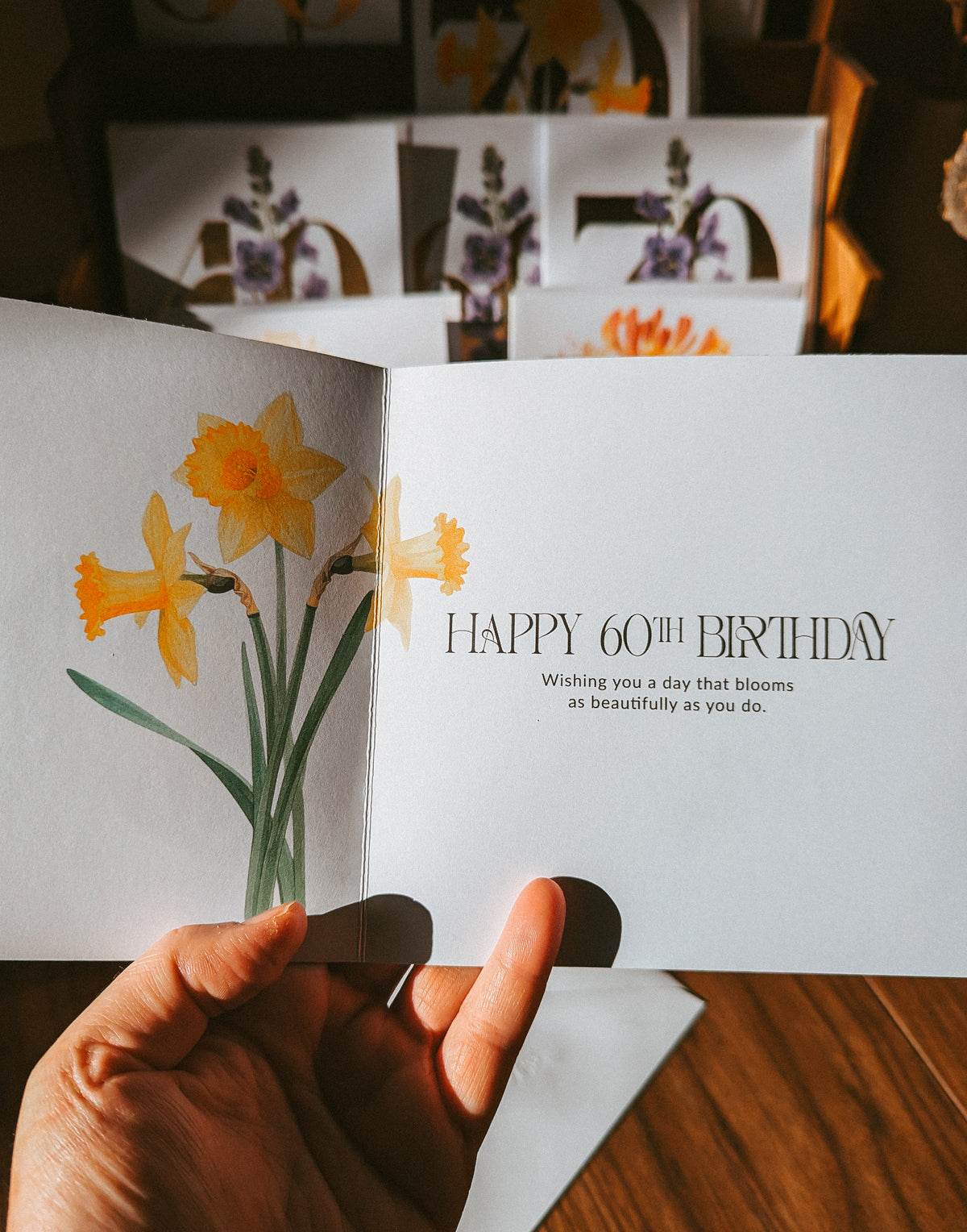 A hand holds an open greeting card featuring yellow daffodils and the text 'HAPPY 60TH BIRTHDAY' on a white card, with more cards stacked in the background on a wooden surface.