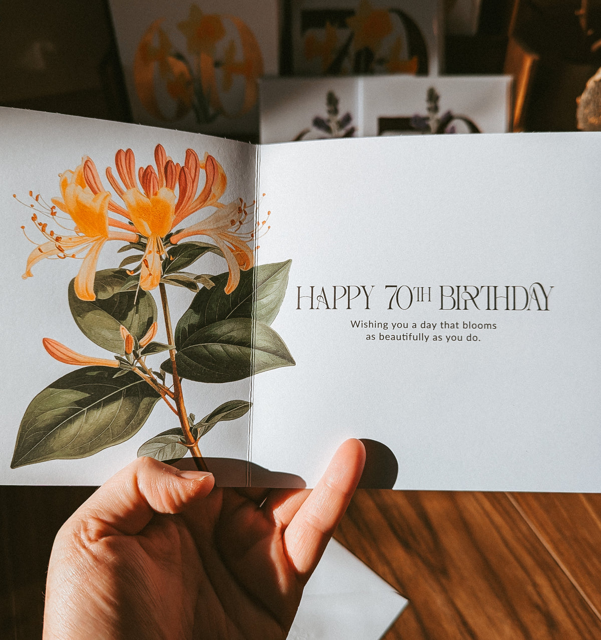 A hand holds an open greeting card with a botanical illustration of orange and yellow flowers on the left page and the text 'HAPPY 70TH BIRTHDAY' on the right page, set against a warm wooden tabletop background.