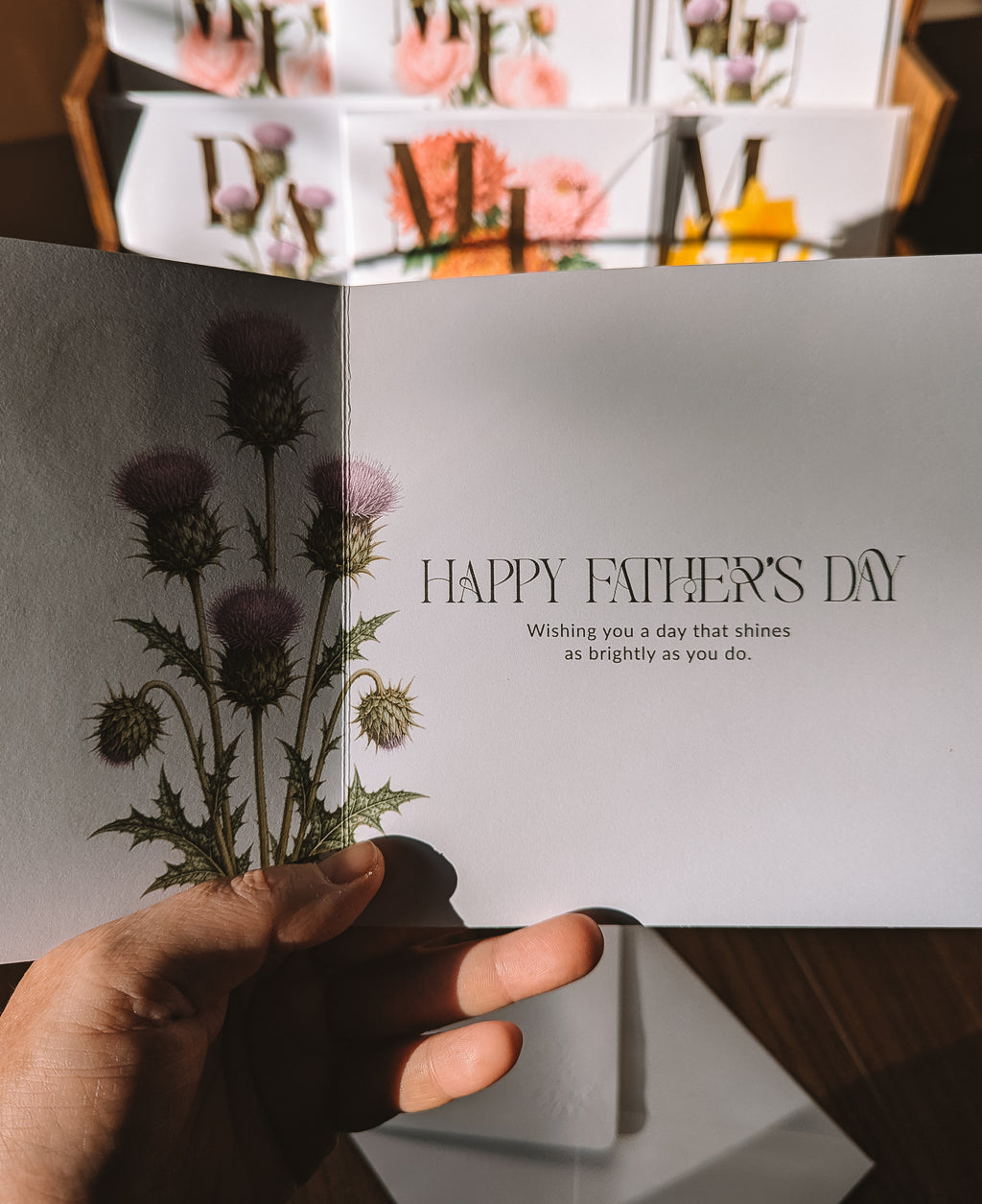 An open Father's Day greeting card held in a hand, featuring a botanical illustration of purple thistles and the printed message 'HAPPY FATHER'S DAY' with a smaller quote below. The background shows a blurred display of similar greeting cards.