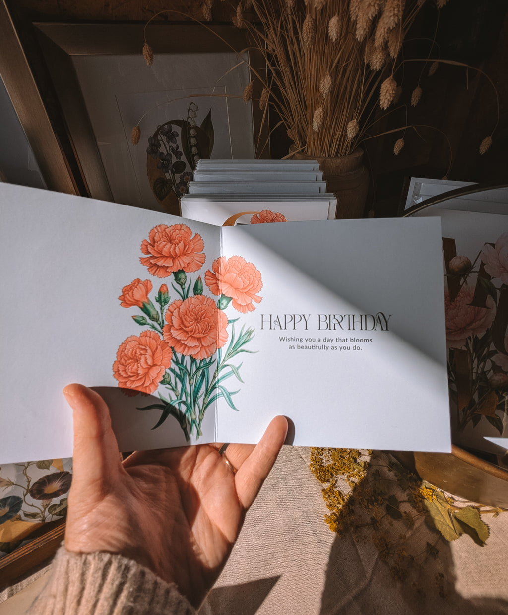 A hand holds an open greeting card with a watercolor-style floral illustration and the text 'HAPPY BIRTHDAY' on a warm, sunlit tabletop surrounded by dried flowers and framed art.