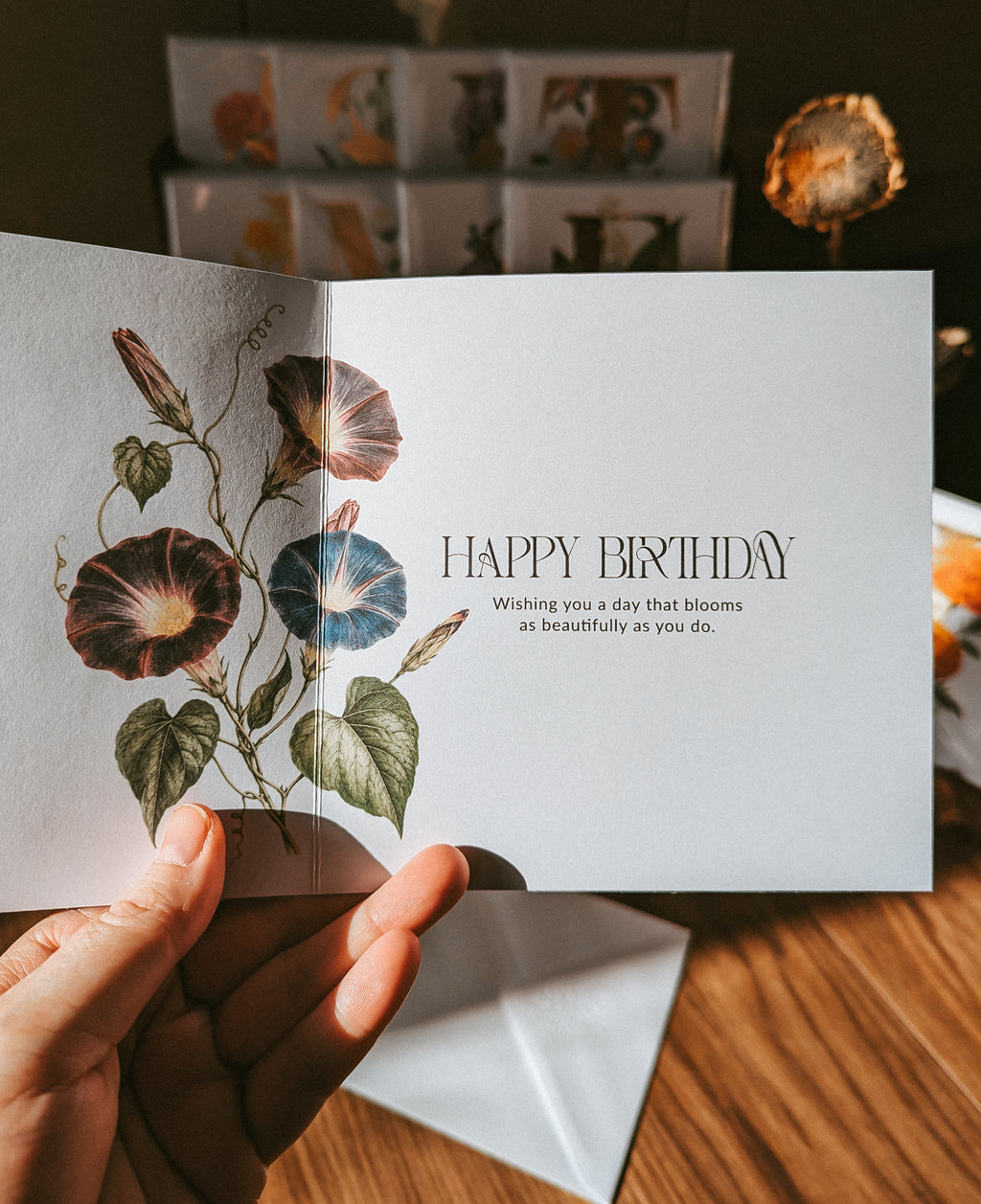 A hand holds an open greeting card with a botanical illustration of flowers on the left panel and the words “HAPPY BIRTHDAY” on the right panel. The card is photographed on a wooden table with other cards and dried flowers blurred in the background.