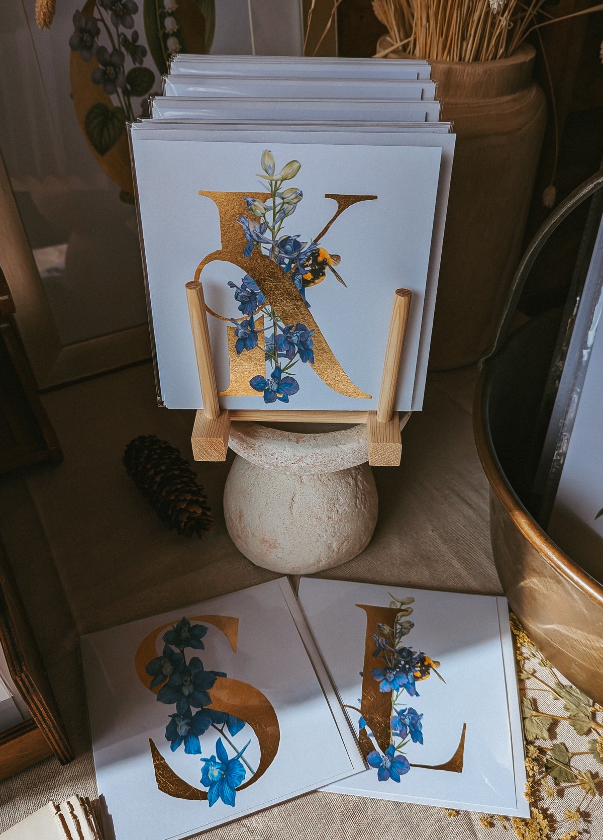 A stack of personalised square cards displayed on a small wooden stand, with two cards laid flat in front showing large gold letters decorated with purple and blue Larkspur flowers. The setting appears styled in a rustic indoor environment with dried florals and neutral decor.
