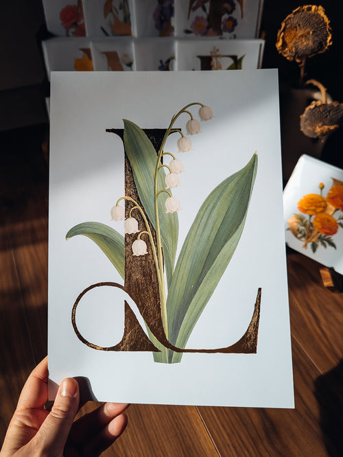 A hand holds a white art print featuring a botanical illustration of a lily of the valley flower integrated into a large decorative letter 'L' on a white background, set against a warm wooden table with dried flowers in the background.