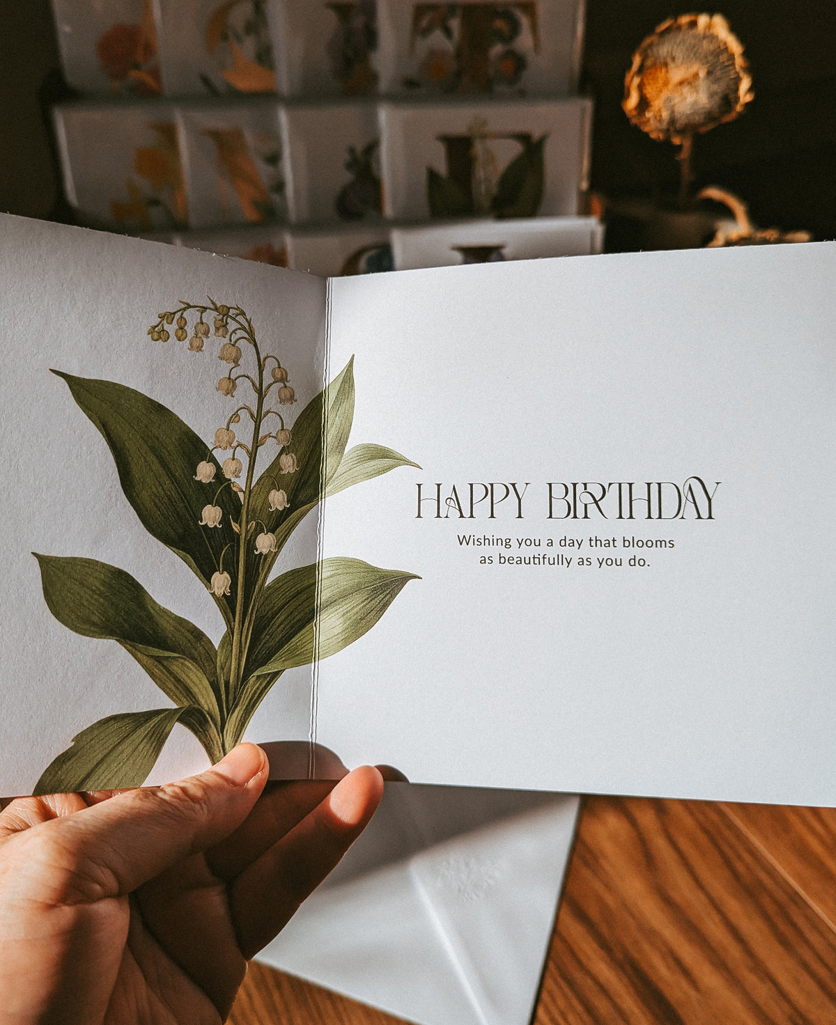 A hand holds an open botanical birthday card featuring a lily of the valley illustration on the left page and the words 'HAPPY BIRTHDAY' on the right page, with a matching envelope and blurred cards in the background.