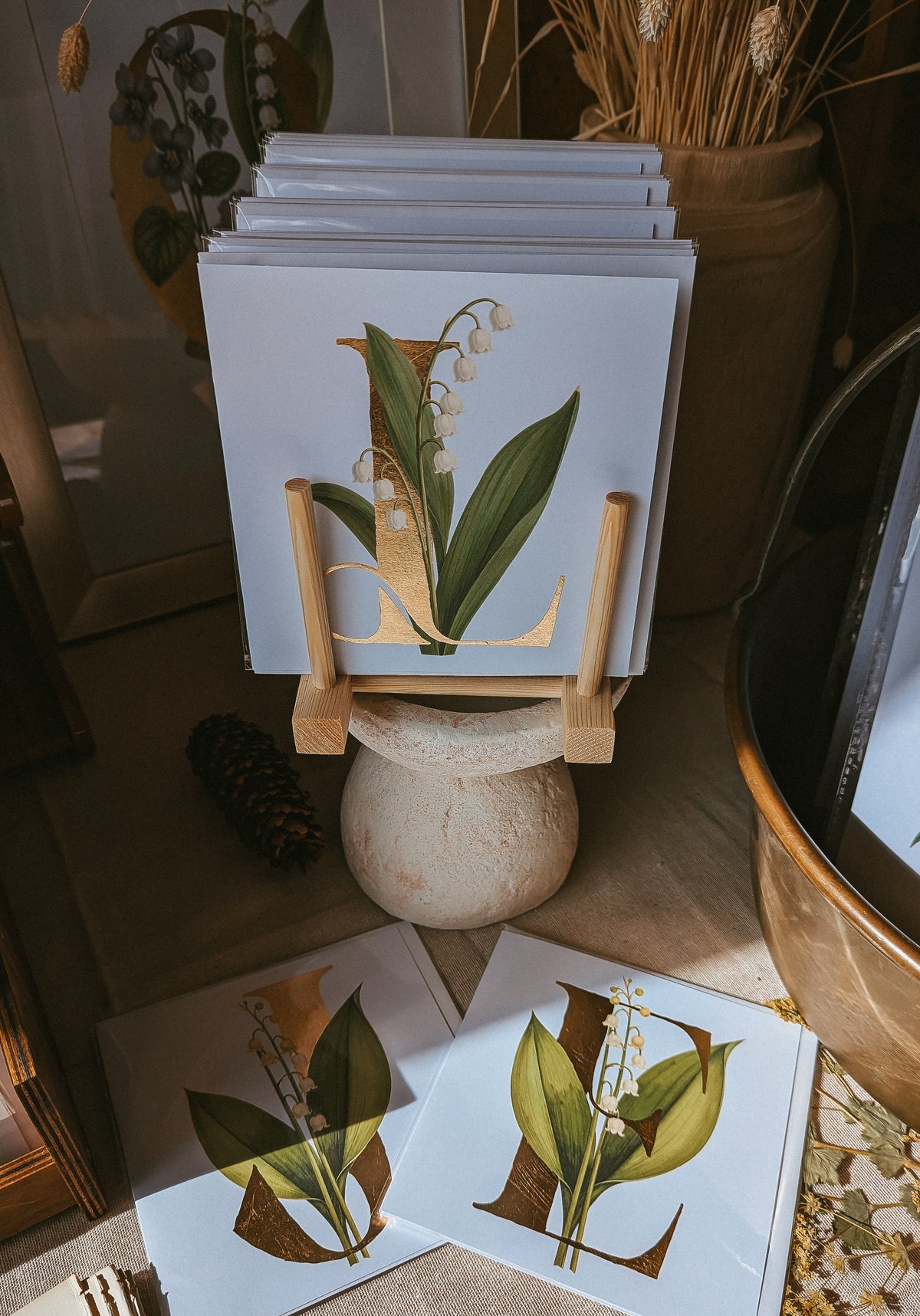A stack of botanical art cards featuring the letter 'L' paired with a white lily of the valley and leaves, displayed on a small wooden stand in a styled interior setting.