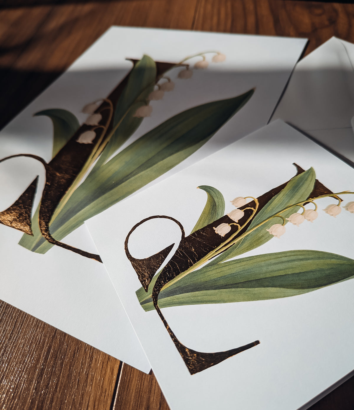 Card & Print laid on a wooden surface, featuring a botanical illustration of a lily-of-the-valley with green leaves and small white flowers on white paper.