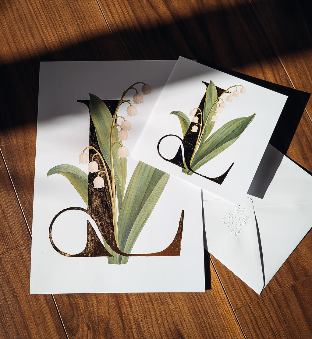 A styled flat-lay of a white greeting card and print featuring a large decorative letter L adorned with lily of the valley flowers and green leaves, shown on a wooden floor with a matching envelope.