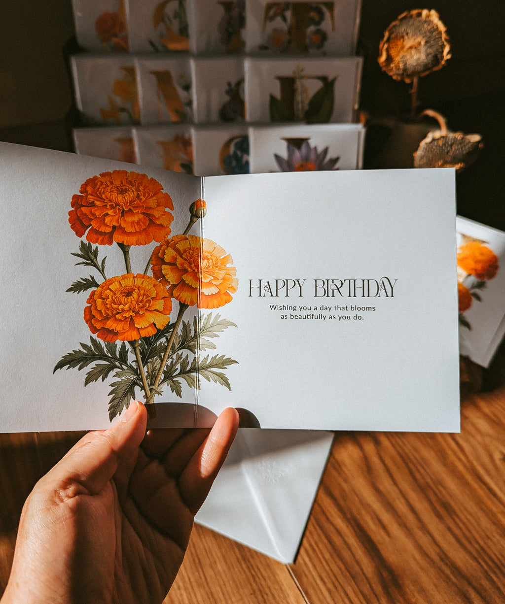 A hand holds an open square greeting card with orange marigold flowers on the left panel and a warm message on the right panel reading “HAPPY BIRTHDAY.” The card is photographed on a wooden surface with stacked floral cards in the background.