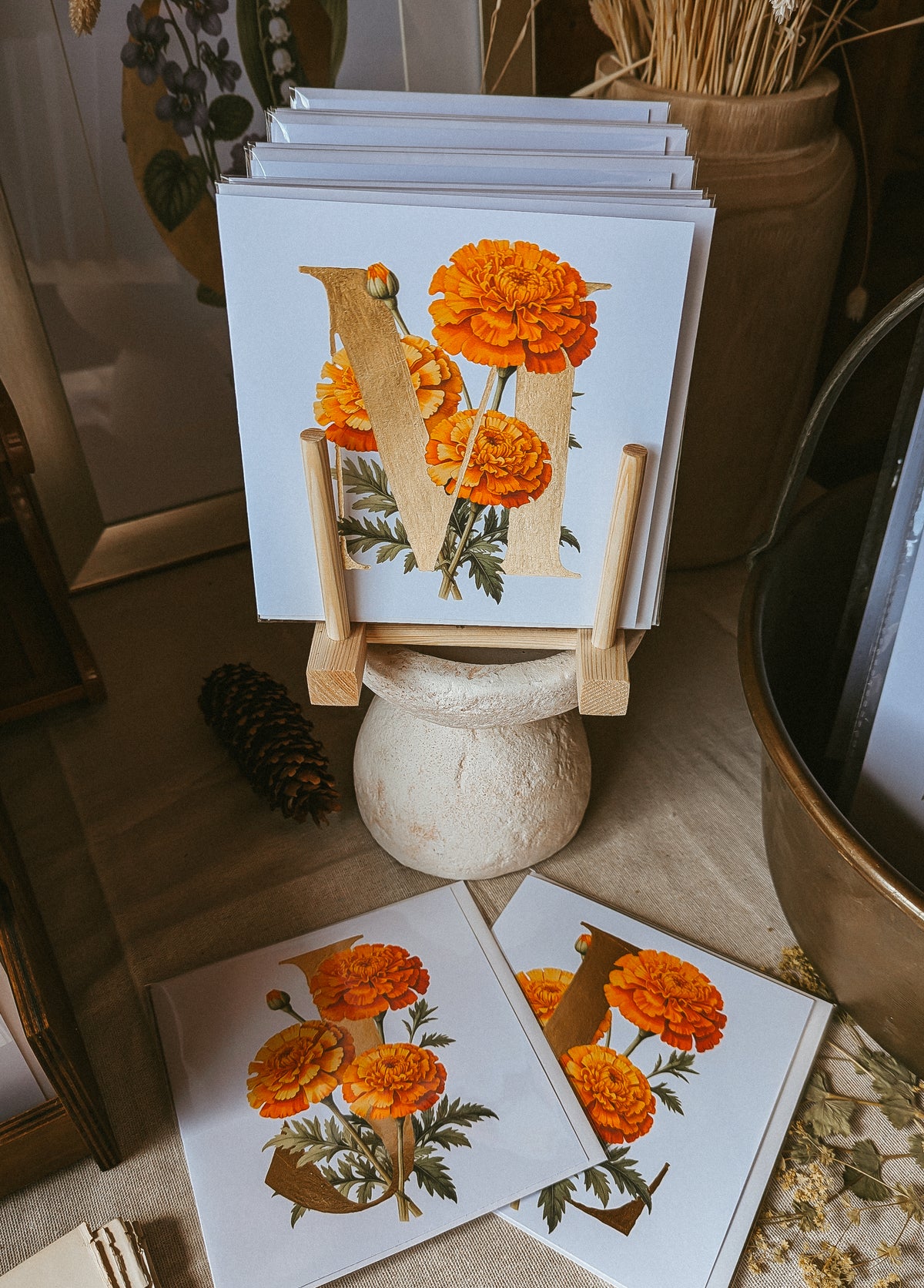 A stack of art cards featuring a large gold letter 'M' decorated with orange marigold flowers and green leaves, displayed on a small wooden stand on a tabletop.