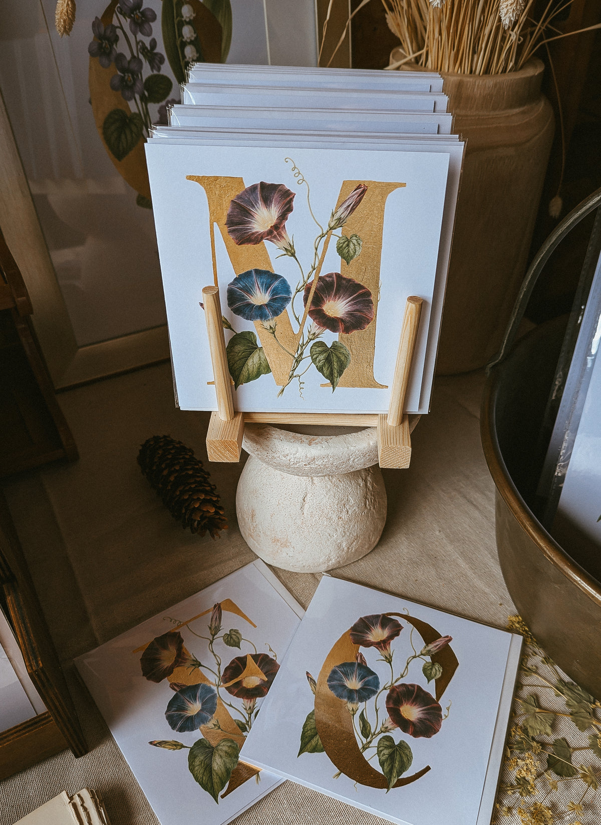 A stack of art cards featuring a large gold foil letter 'M' decorated with botanical illustrations of morning glory flowers, displayed on a small wooden stand in a styled home decor setting.