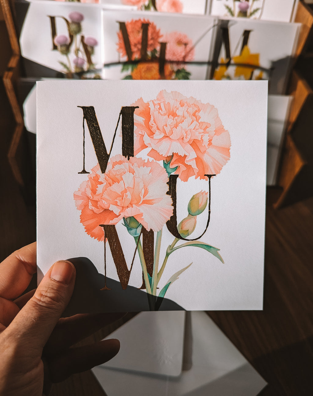 A hand holds a square card featuring the word 'MUM' in large gold foil serif letters overlaid with soft peach carnation flowers and green leaves on a white background. The scene is indoors with blurred floral prints and wooden furniture in the background.