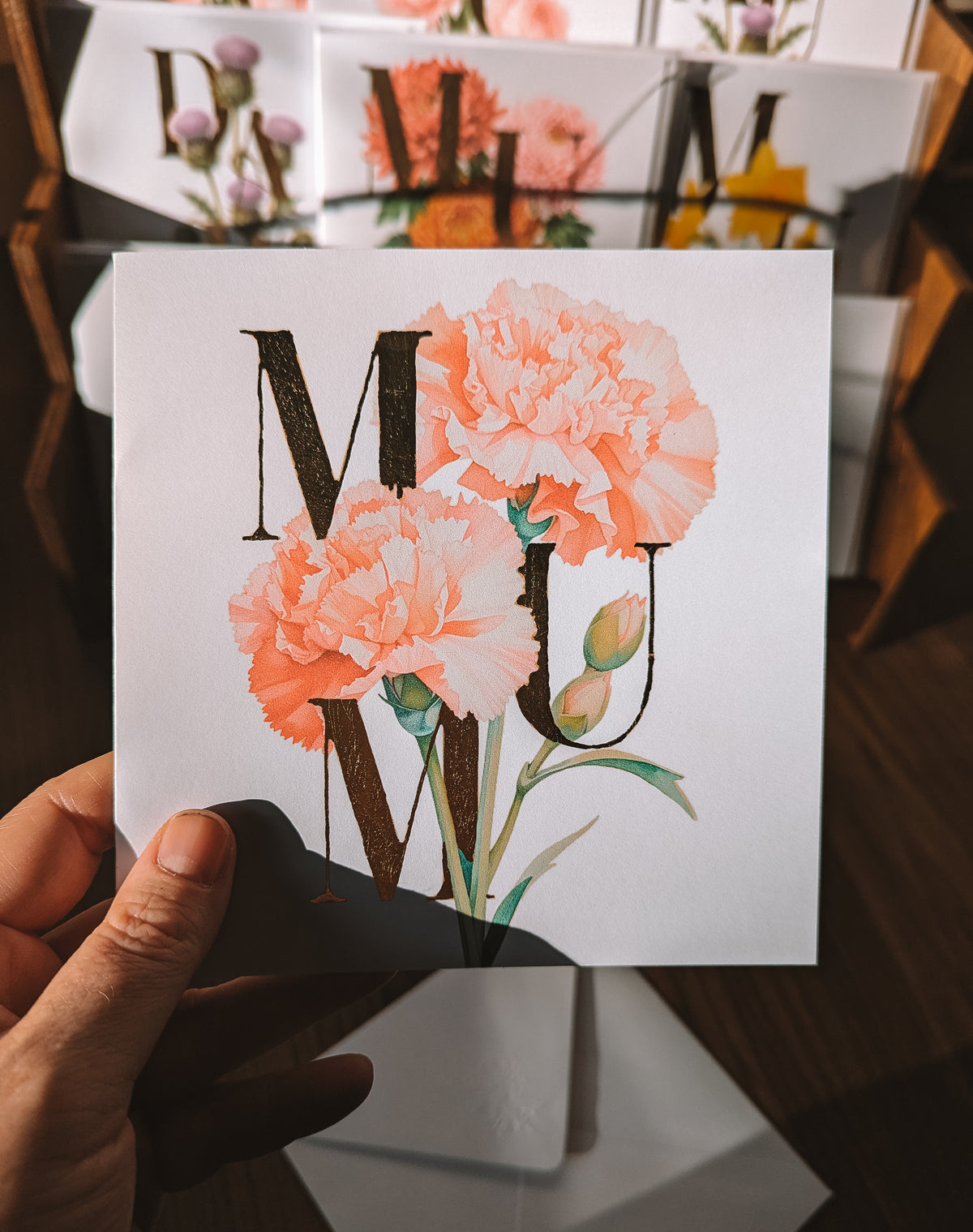 A hand holds a square card featuring the word 'MUM' in large gold foil serif letters overlaid with soft peach carnation flowers and green leaves on a white background. The scene is indoors with blurred floral prints and wooden furniture in the background.