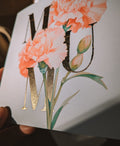A close-up of a printed card featuring large peach and white carnation flowers with green leaves and stems, overlaid with gold foil decorative lettering. A hand is holding the card against a dark background.