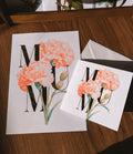 A square white greeting card and matching print on a wooden surface. The card features large gold foil letters 'MUM' decorated with watercolor-style peach and pink carnation flowers with green stems.
