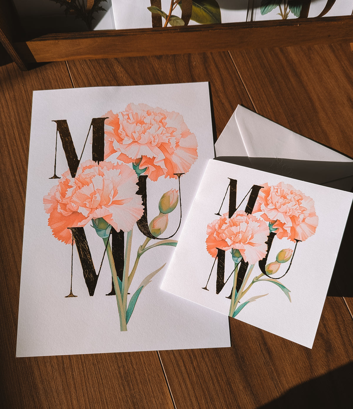 A square white greeting card and matching print on a wooden surface. The card features large gold foil letters 'MUM' decorated with watercolor-style peach and pink carnation flowers with green stems.