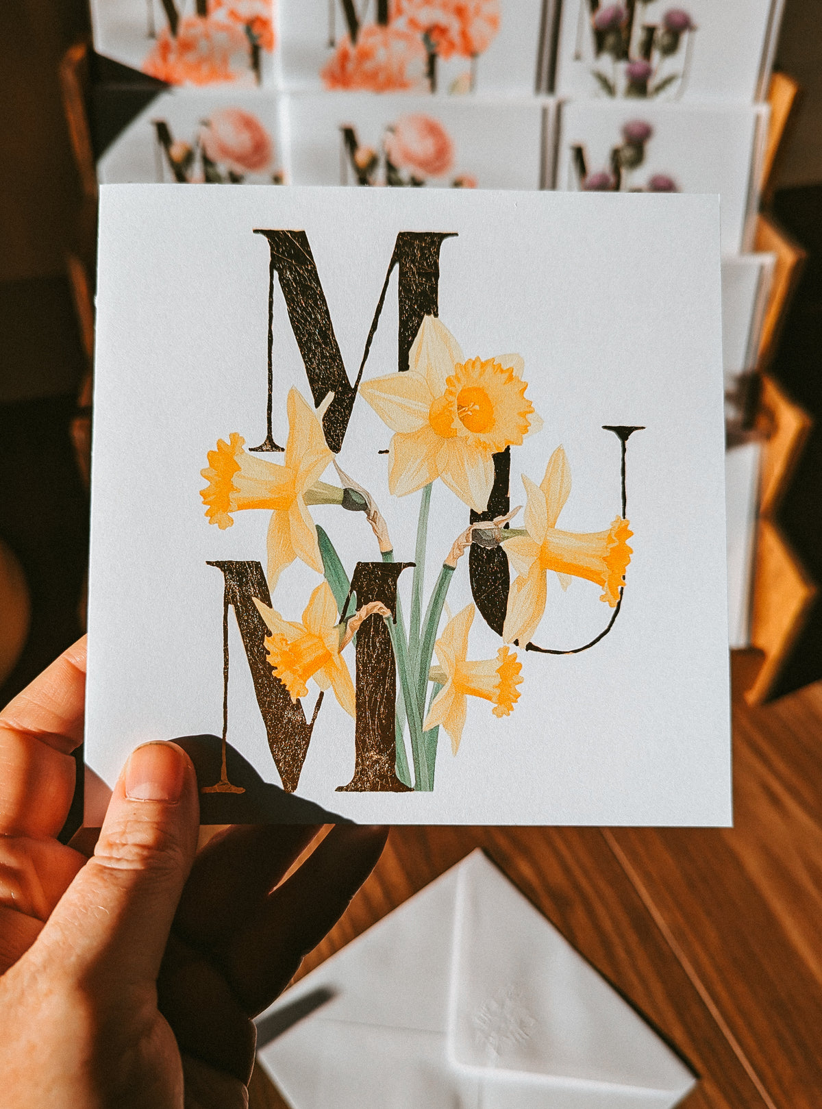 A hand holds a square white greeting card featuring large gold foil letters 'MUM' decorated with yellow daffodils and green stems. The background shows a wooden table and more matching cards.