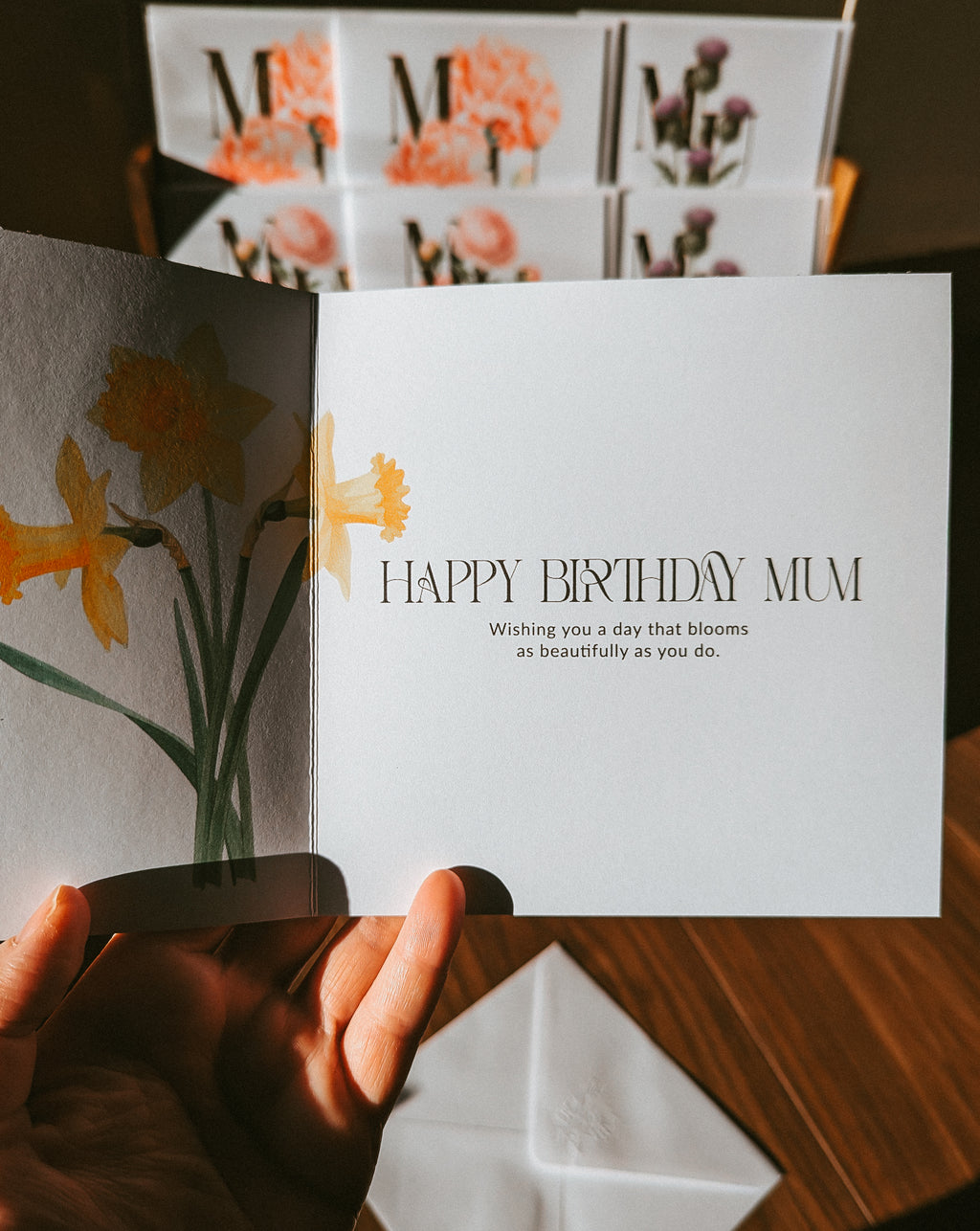 A hand holds an open greeting card with a watercolor daffodil design on the left panel and the text 'HAPPY BIRTHDAY MUM' on the right panel. A matching folded card and a white envelope sit on a wooden surface in the foreground, with more floral greeting cards blurred in the background.