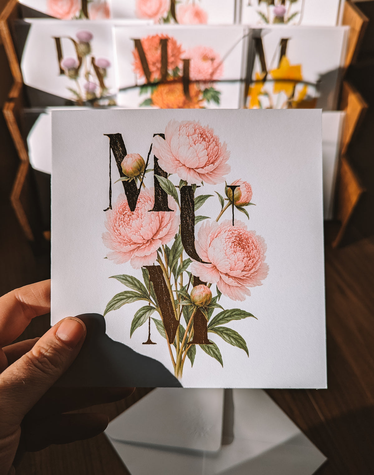 A hand holds a square white card featuring pink peony flowers and large decorative gold foil letters, with blurred matching prints in the background on a wooden surface.