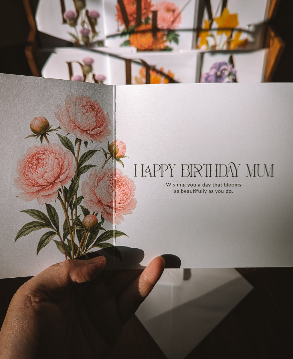 A hand holds an open greeting card with a botanical illustration of pink peonies on the left page and the text 'HAPPY BIRTHDAY MUM' on the right. The background shows more floral greeting cards in a blurred display.