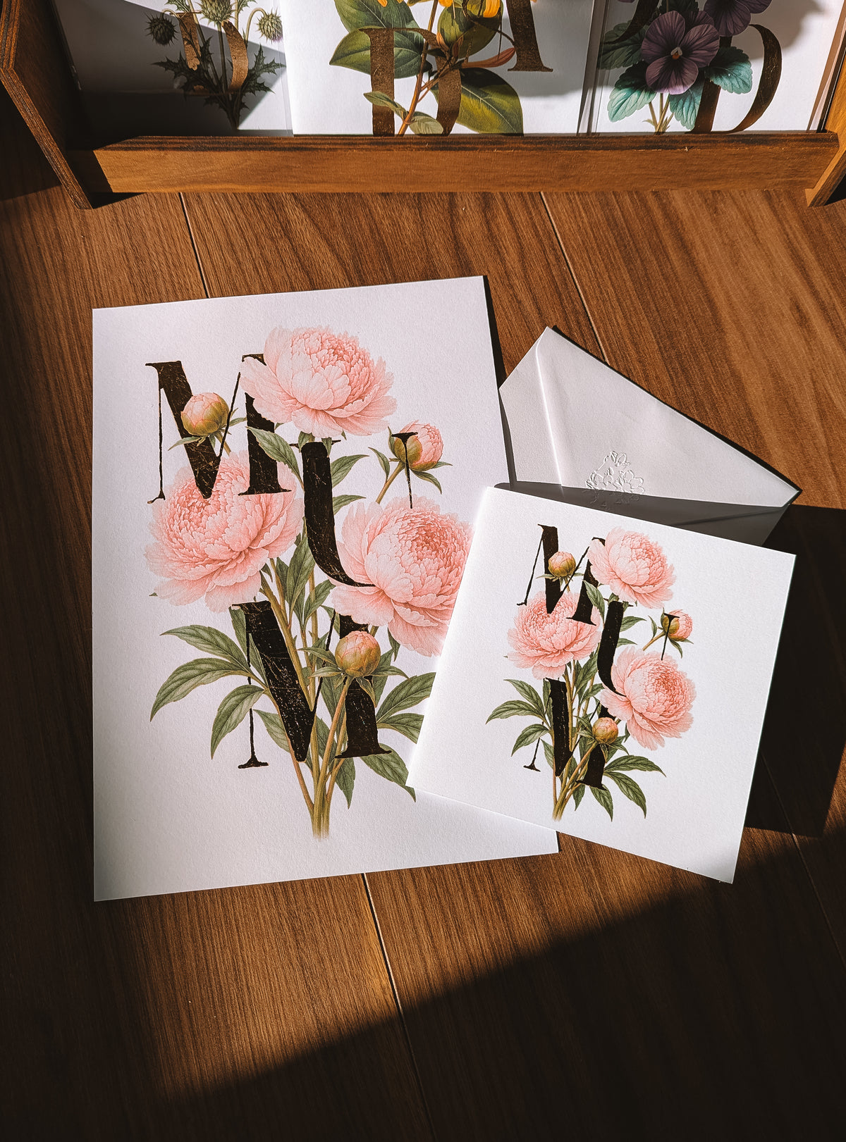 White greeting card & Print with a botanical floral design featuring pink peonies and gold foil decorative letters, displayed on a wooden table with one card open to reveal a white envelope.