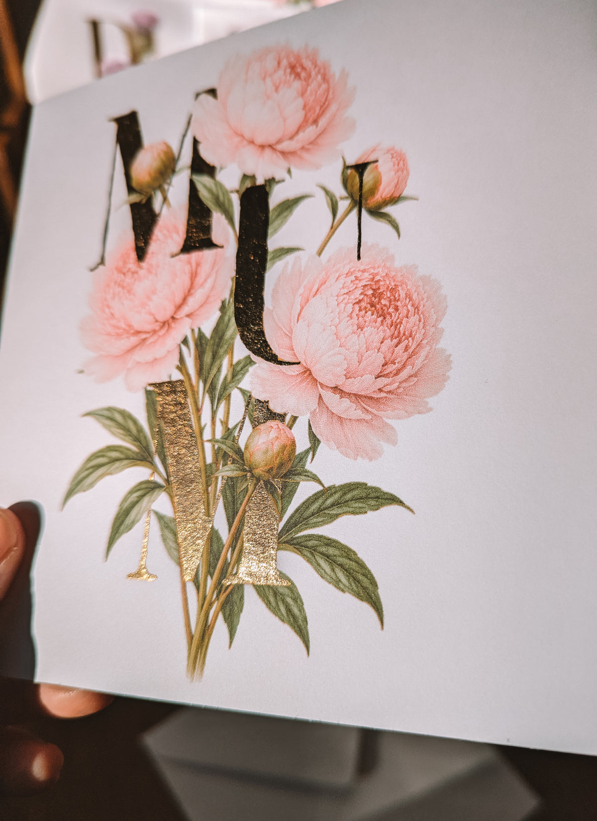 A close-up of a printed card featuring pink peony flowers, green leaves, and large gold foil decorative letters on a white background. A hand is holding the card.
