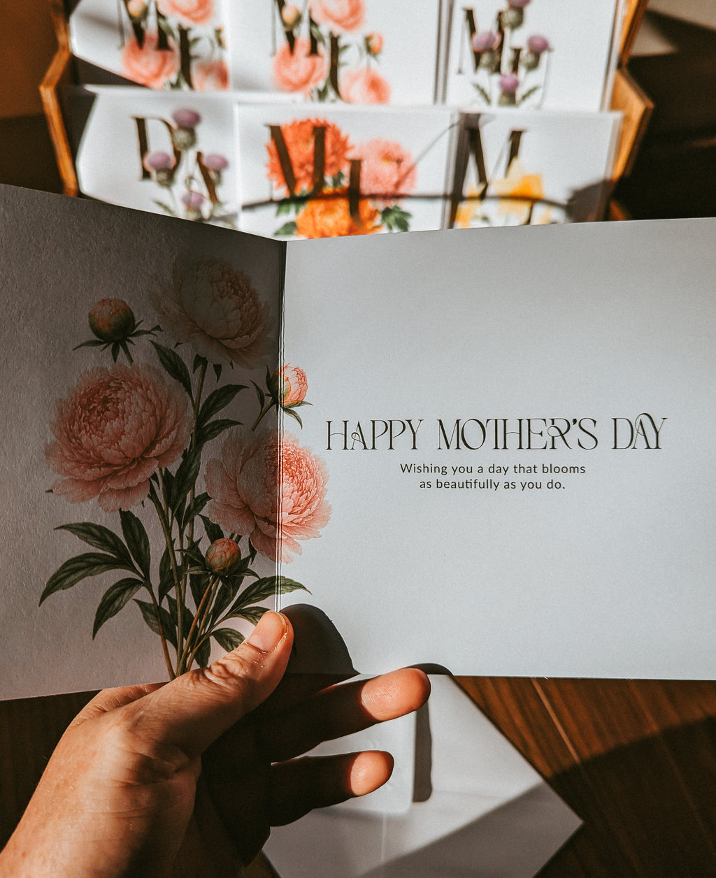 A hand holds an open Mother's Day greeting card with a floral illustration on the left page and the text 'HAPPY MOTHER'S DAY' on the right. More matching cards are stacked in the background on a wooden surface.