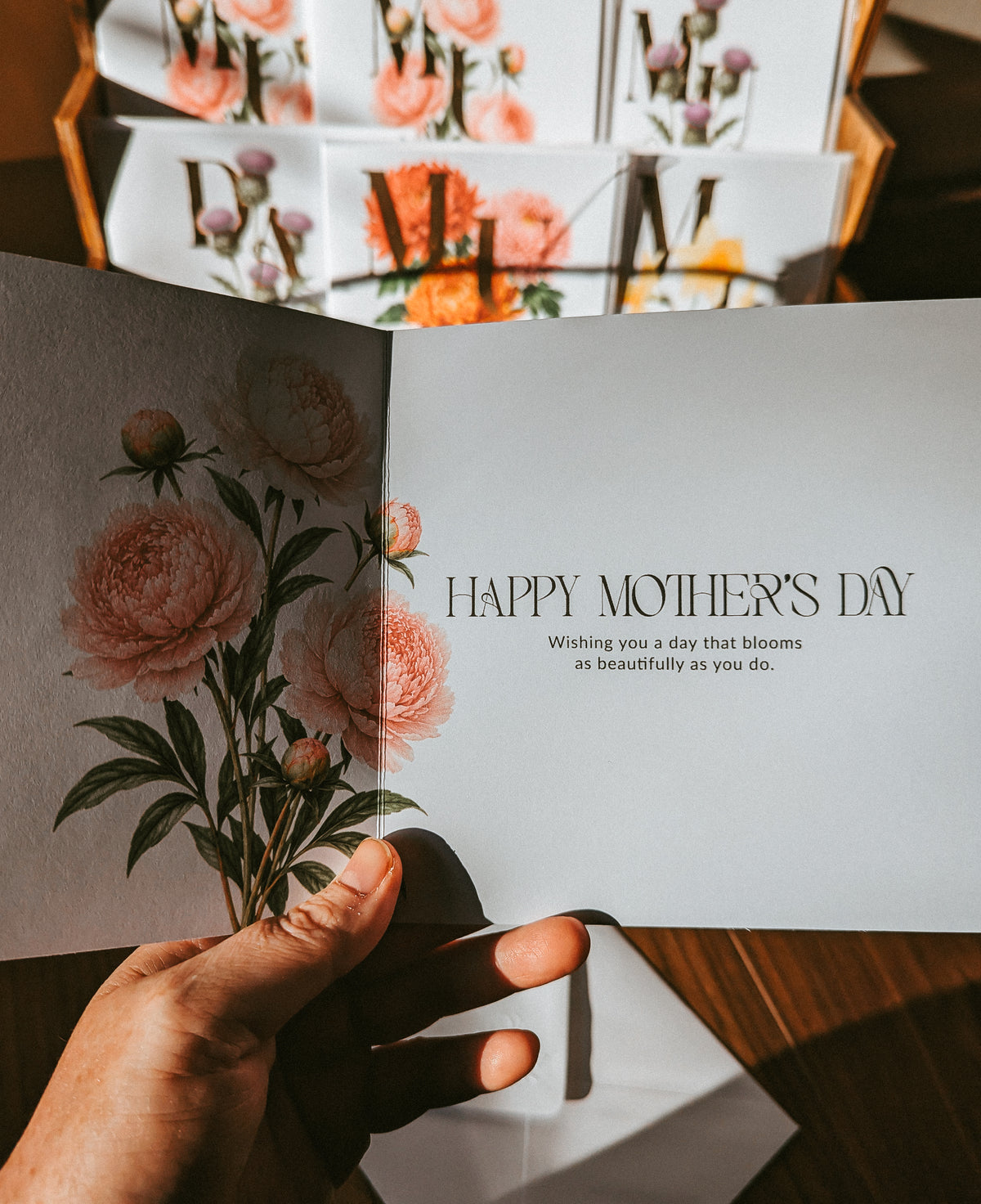A hand holds an open Mother's Day greeting card with a floral illustration on the left page and the text 'HAPPY MOTHER'S DAY' on the right. More matching cards are stacked in the background on a wooden surface.