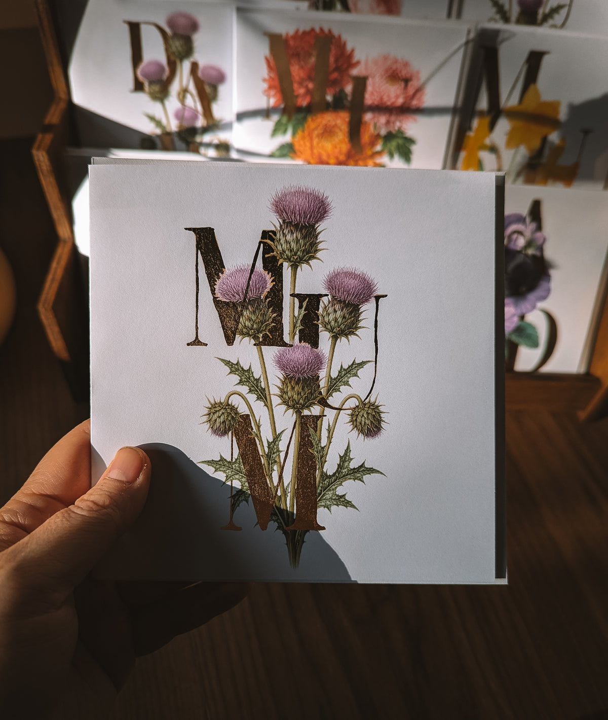 A hand holds a white greeting card featuring a vintage-style floral design with purple thistles and large decorative gold foil letters. The background shows a wooden floor and other cards with floral artwork.