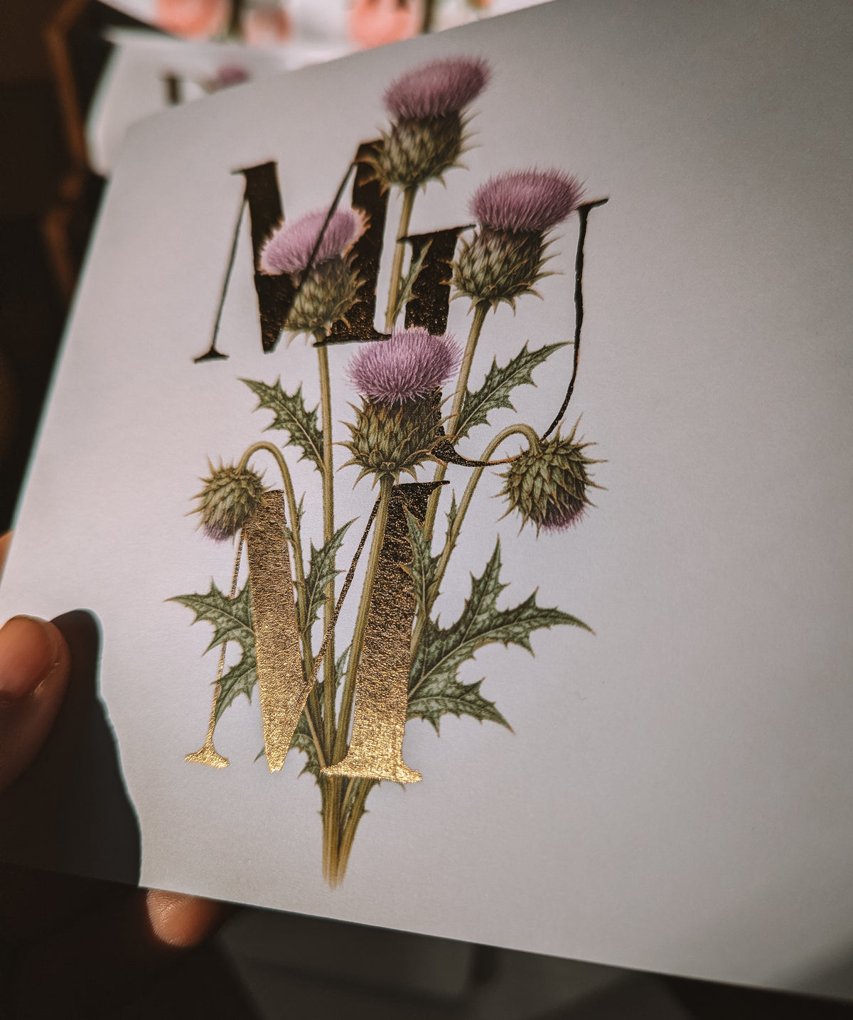 A close-up of a printed card featuring a botanical design with purple thistle flowers, green spiky leaves, and large decorative letters in gold foil, held by a hand against a dark background.
