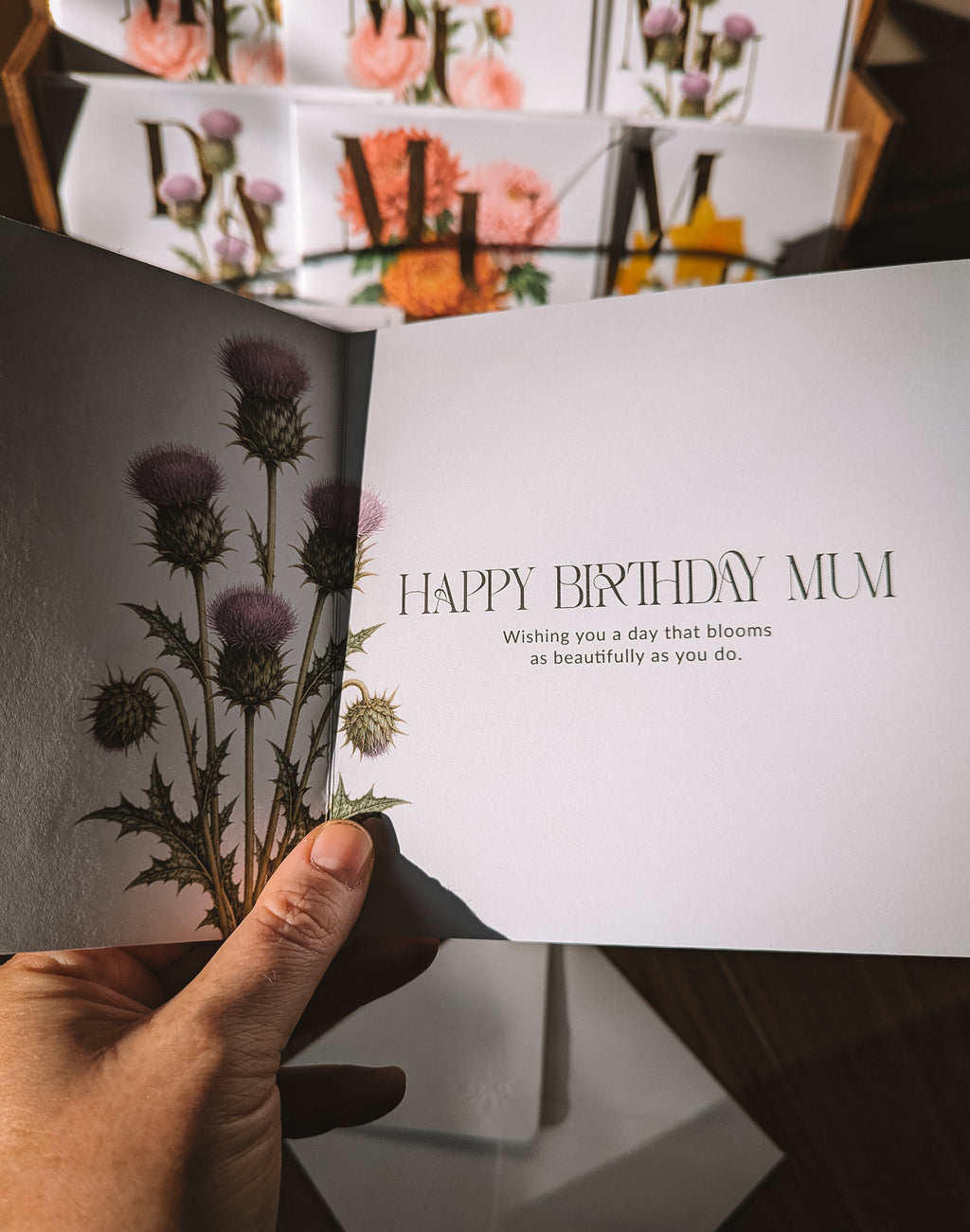 A hand holds an open greeting card with a botanical design of purple thistles on the left panel and the text 'HAPPY BIRTHDAY MUM' on the right panel. The background shows a display of similar floral greeting cards.