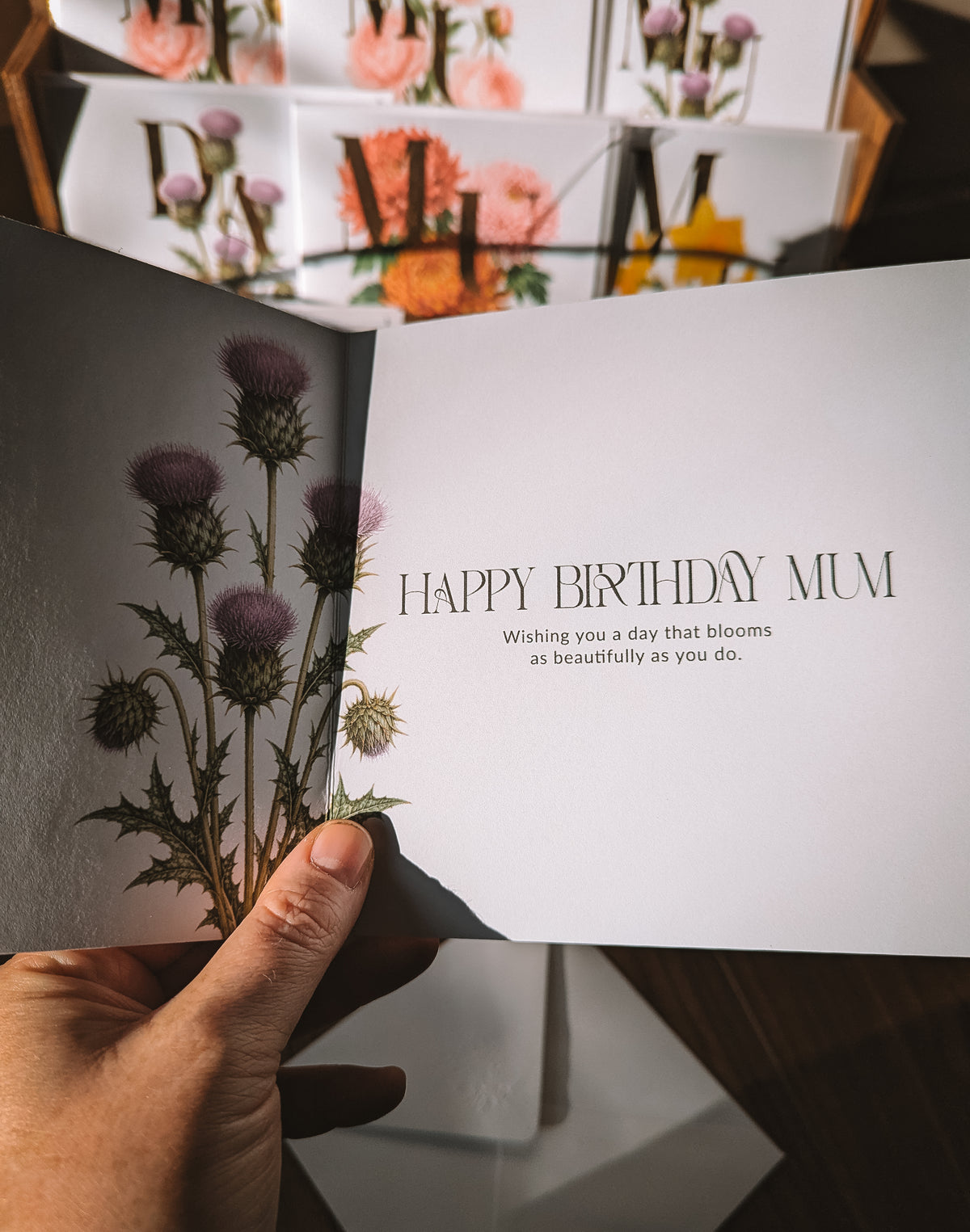 A hand holds an open greeting card with a botanical design of purple thistles on the left panel and the text 'HAPPY BIRTHDAY MUM' on the right panel. The background shows a display of similar floral greeting cards.
