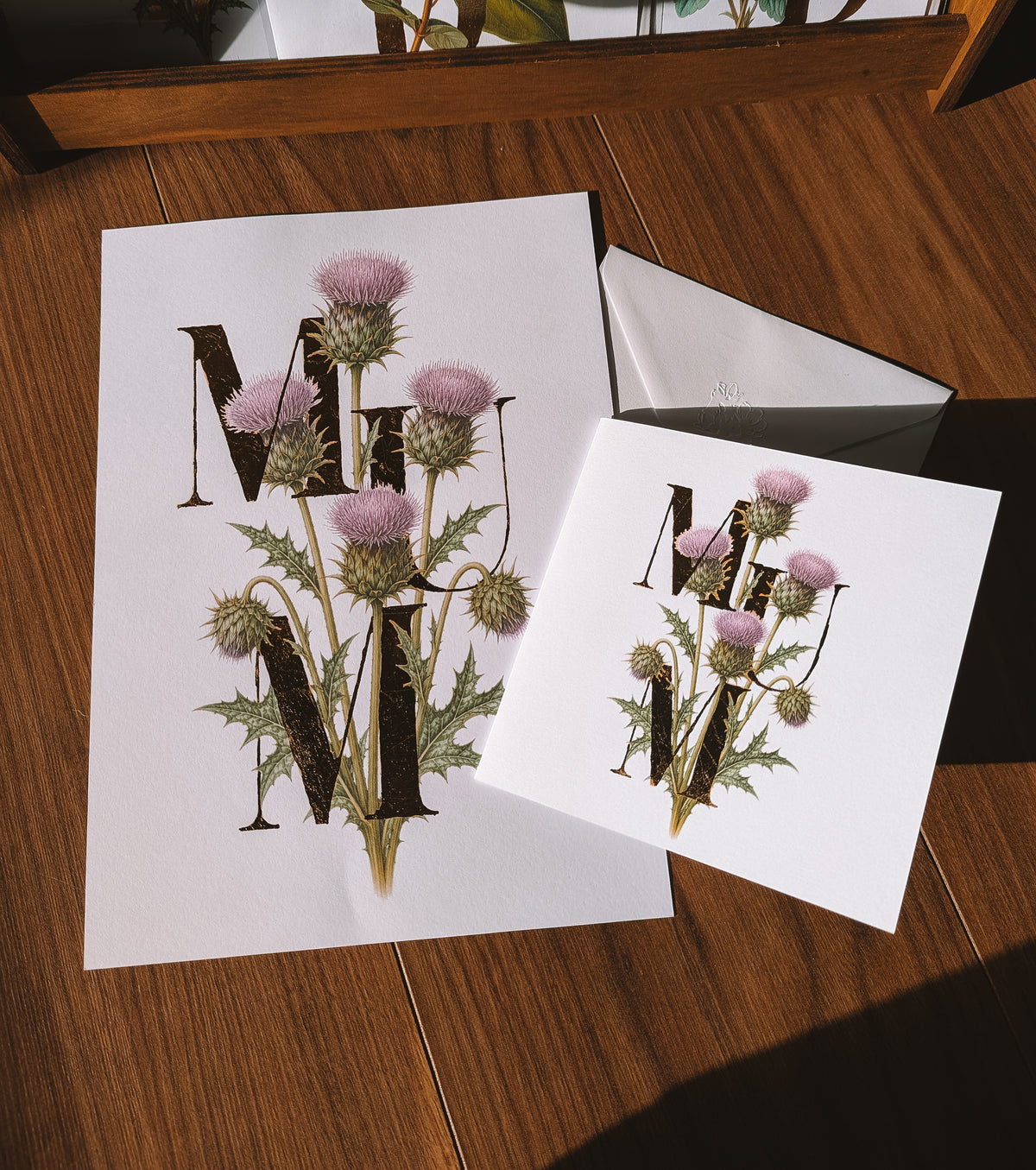 A white greeting card and matching print on a wooden tabletop. The card features a large decorative gold foil letters 'MUM' blended with an illustration of thistle flowers.